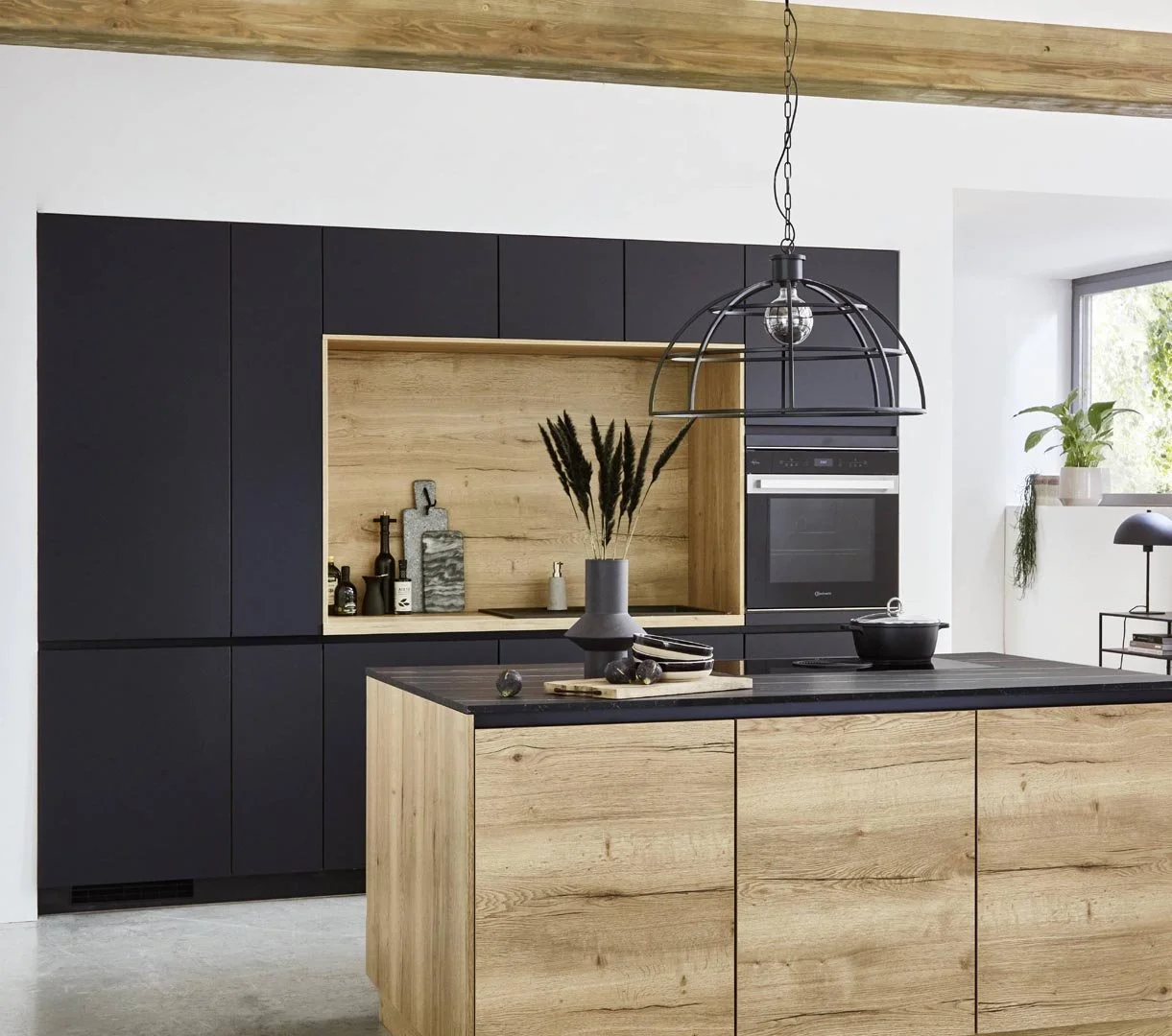 Modern kitchen with black and wooden cabinetry, pendant light, decorative vases and plants, and a window with a view of greenery.
