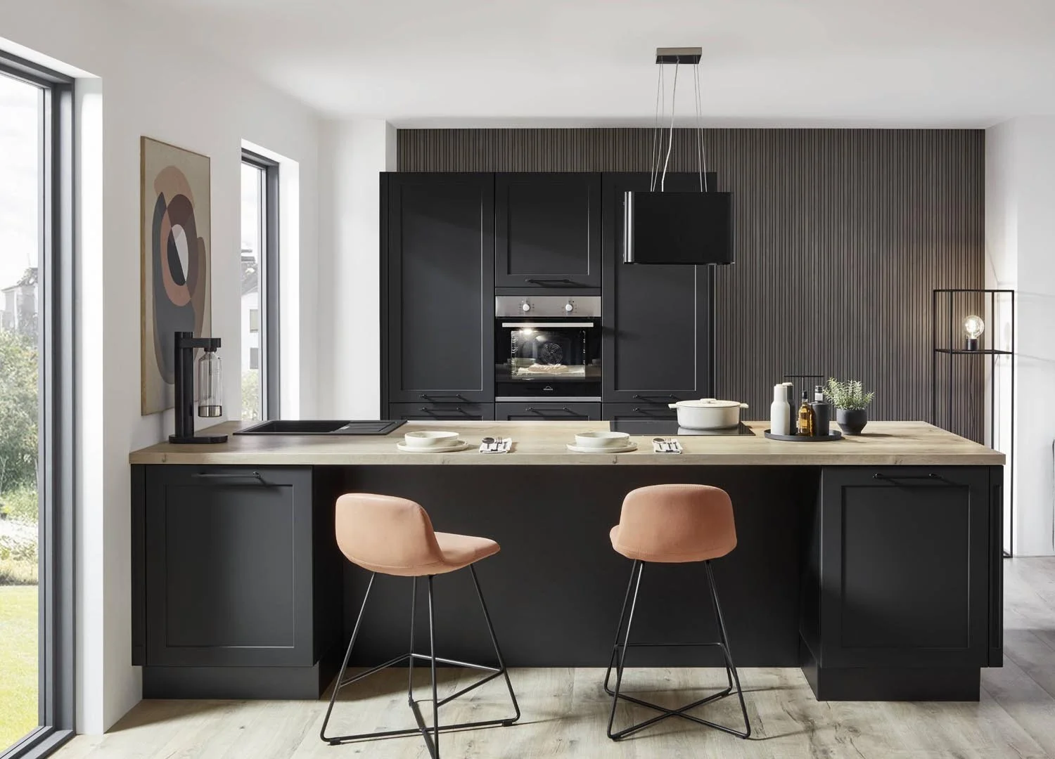 Modern kitchen with black cabinets, a light wooden island, two pink bar stools, a small black tray with bottles and plant on the island, and large windows with a view of greenery.