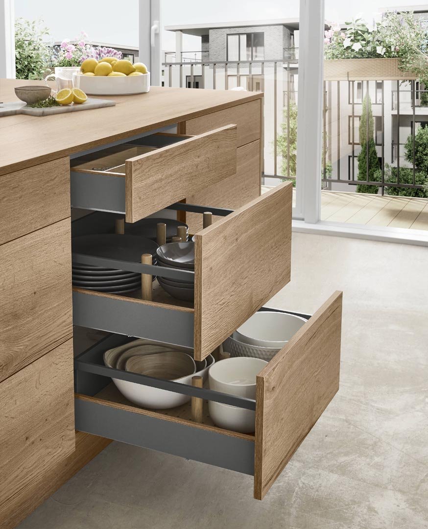 Open kitchen drawers filled with plates and bowls, with a view of a sunlit balcony with potted plants outside.