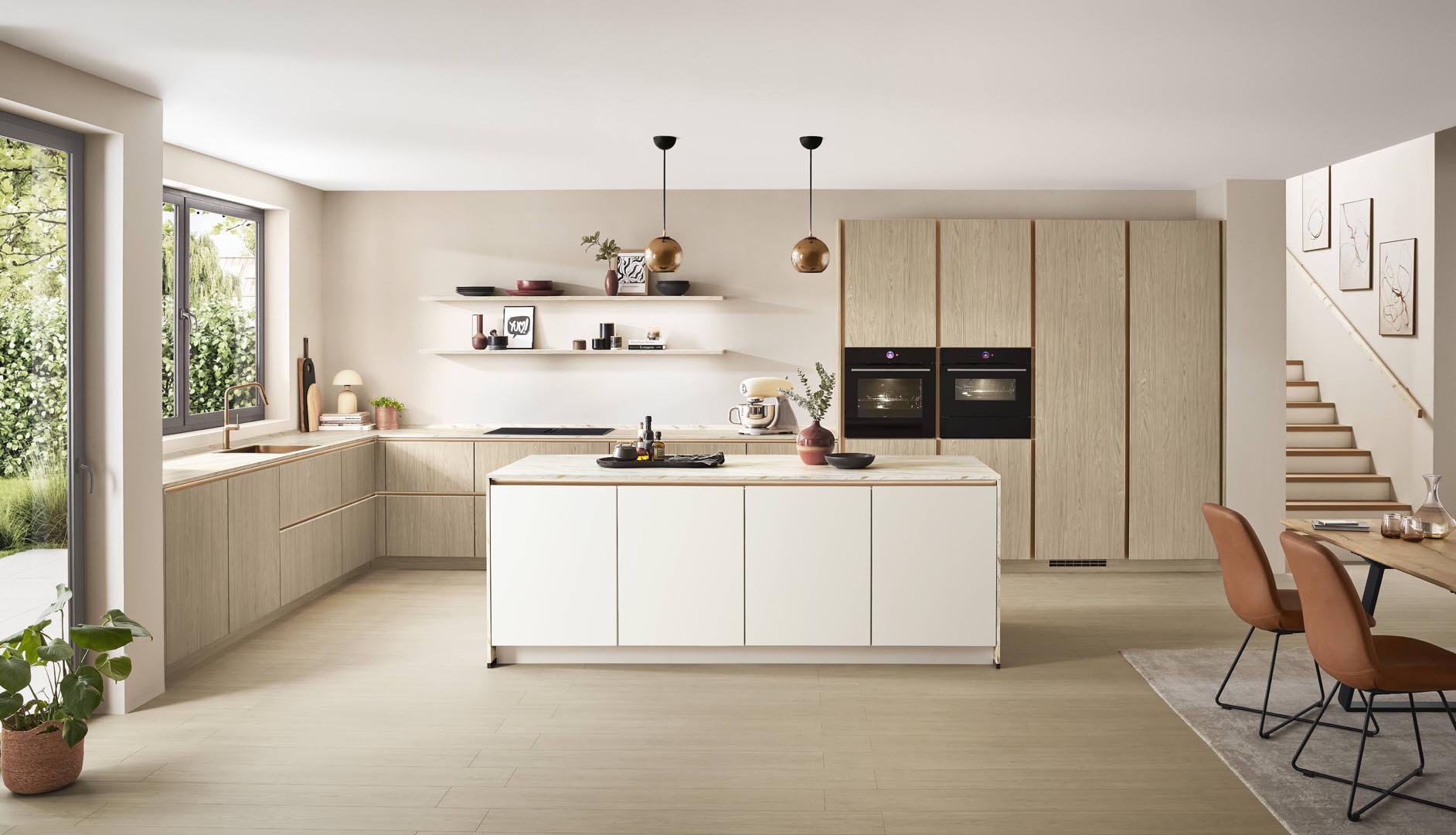 Modern kitchen with light wood cabinetry, white island, two hanging copper pendant lights, large window, open shelving, dining area with terracotta chairs, and staircase with framed artwork.