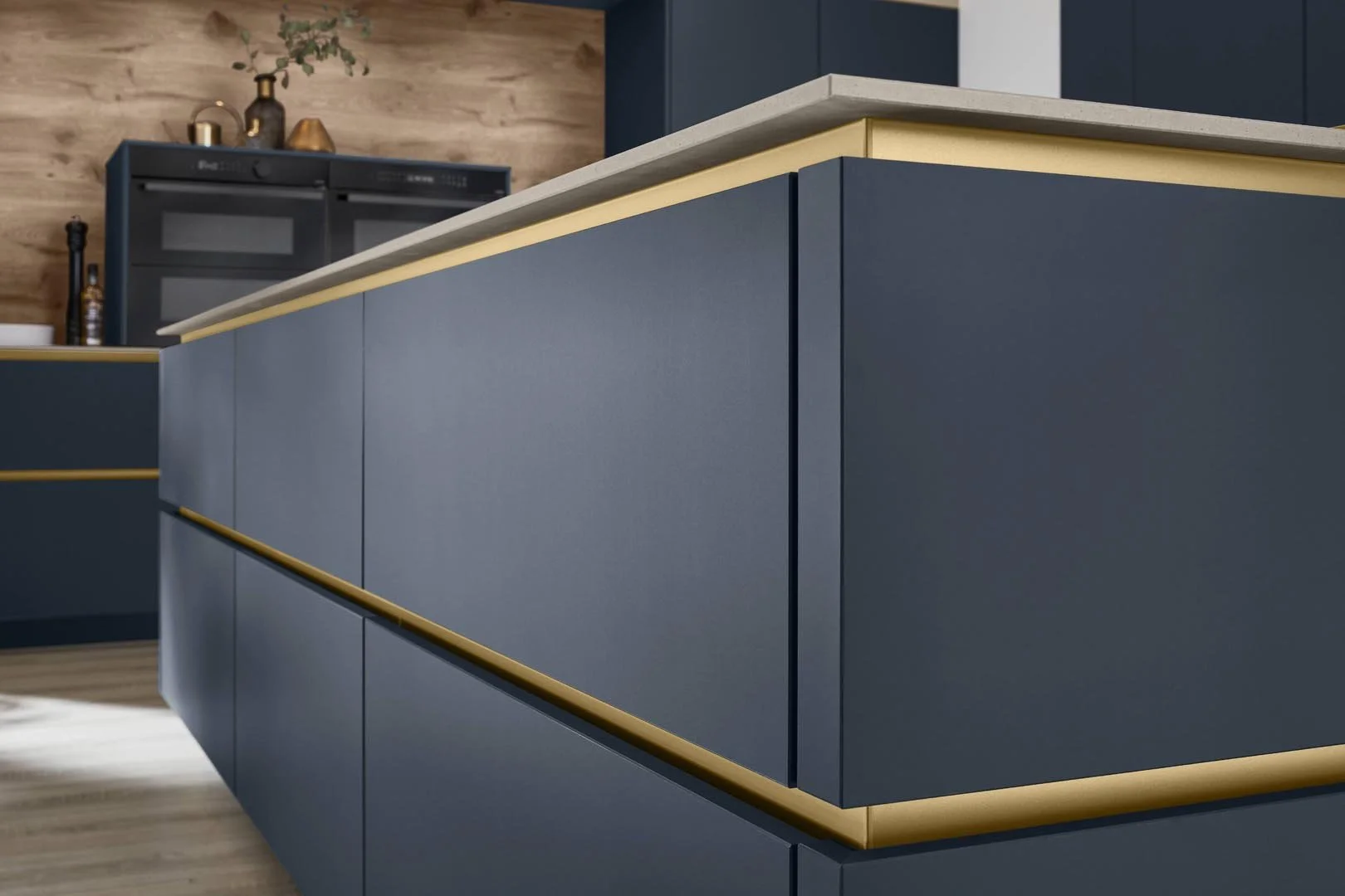 Close-up view of modern kitchen island with blue cabinetry, beige countertop, and gold accents, with wooden wall and kitchen appliances in the background.