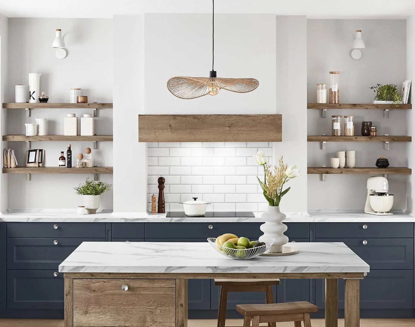 Modern kitchen with white walls, wooden open shelves holding containers, a marble countertop, a navy blue lower cabinet, a white brick backsplash, a hanging wicker light, a white stand mixer, and a central island with a floral arrangement and green a