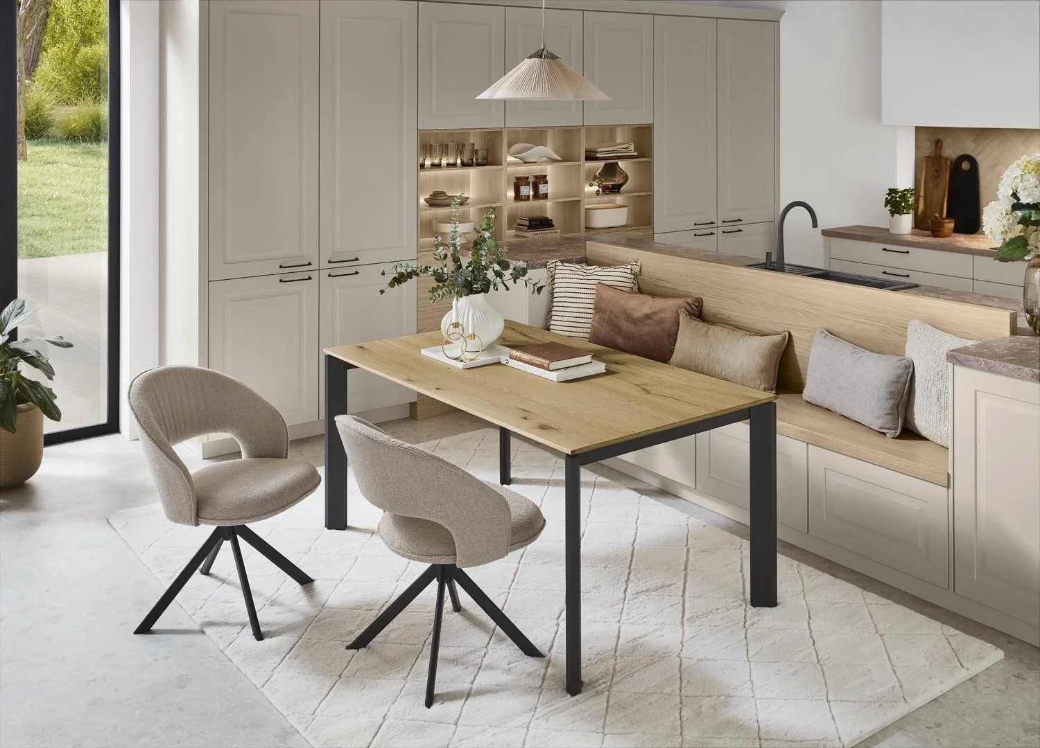 Modern dining area with a wooden table, two beige upholstered chairs, and a built-in bench with pillows, in a room with white cabinetry, open shelves, and a black faucet near a window with a view of a green backyard.
