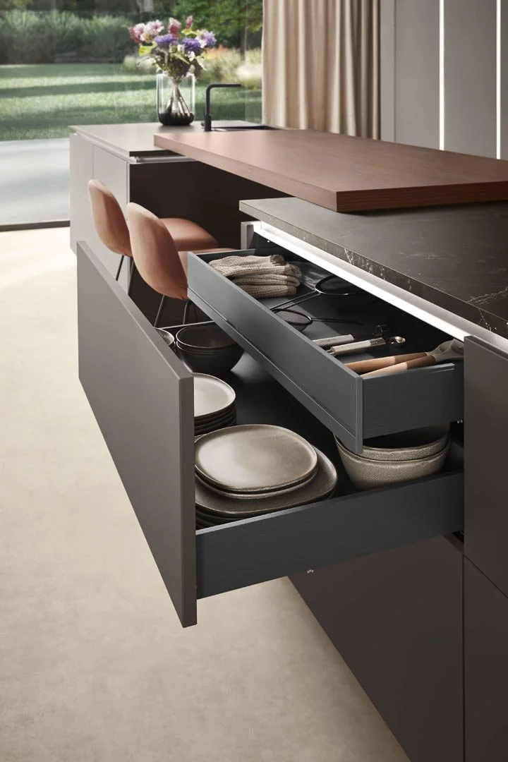 Open kitchen drawer with plates, bowls, and utensils, next to a countertop with a vase of flowers and two peach-colored chairs nearby.