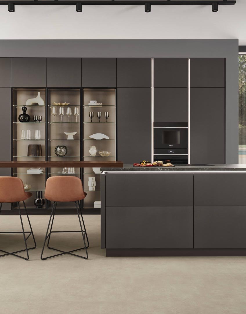 Modern kitchen with dark gray cabinetry, glass shelves with dishes and decorative items, a built-in microwave, and a kitchen island with a black marble countertop. Two peach-colored bar stools are in front of the island, and there are snacks on the c