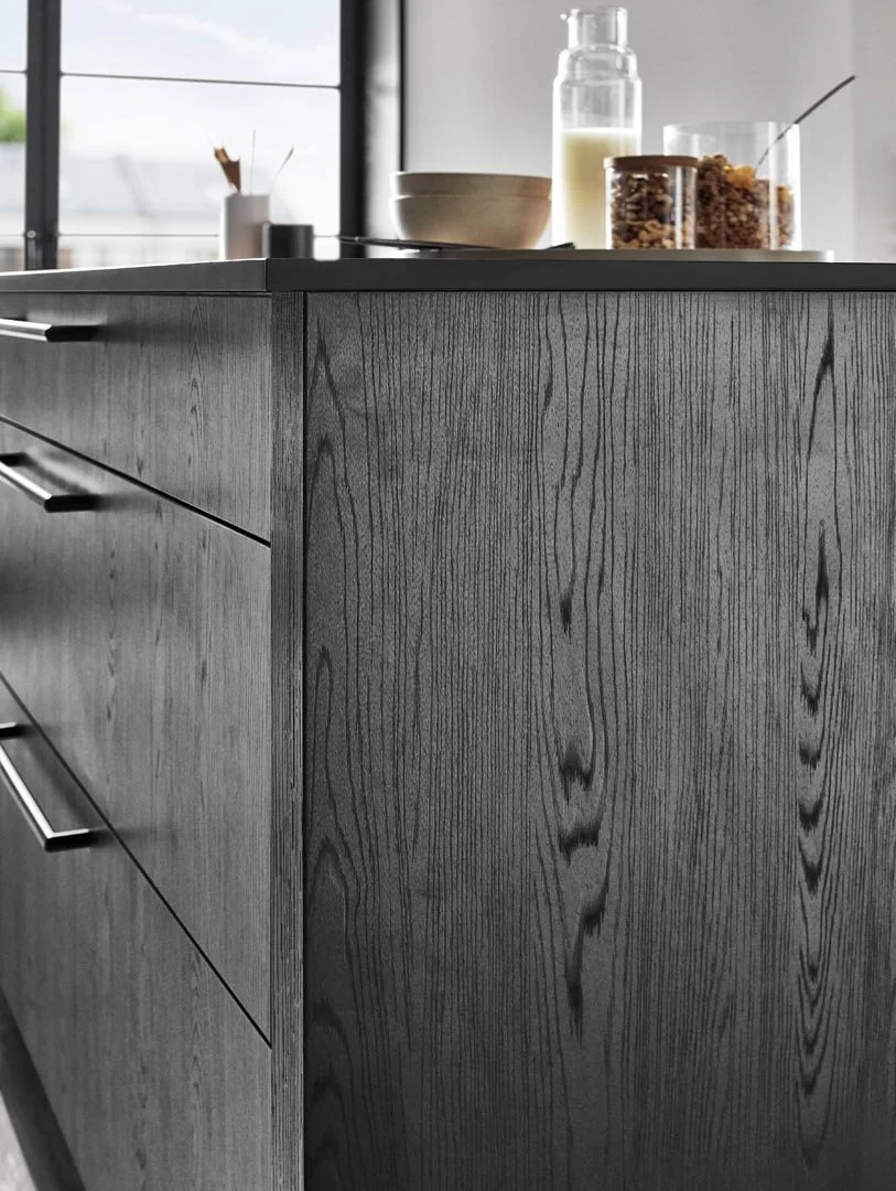 Close-up of a modern kitchen island made of dark wood grain with metal handles, dishes and condiments on the countertop, and a window in the background.