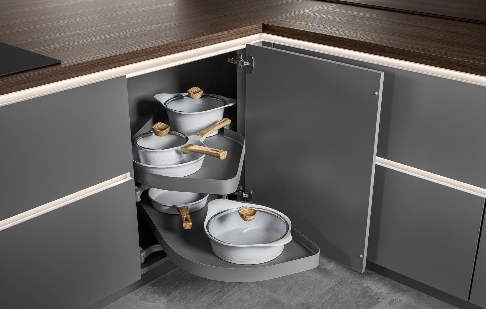 Gray kitchen cabinet with open corner pull-out storage containing white enamel pots with wooden handles and lids.