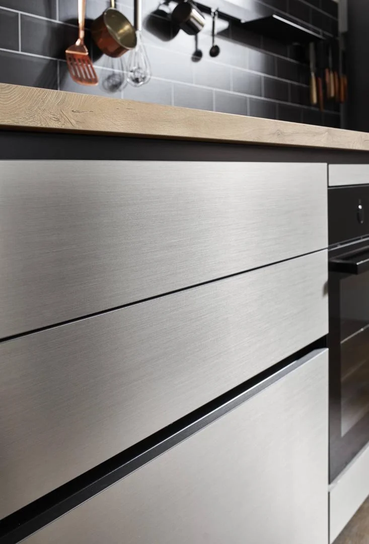 Close-up of modern kitchen drawers with brushed metal finish and wooden countertop, with hanging pots and utensils on a dark tiled backsplash.