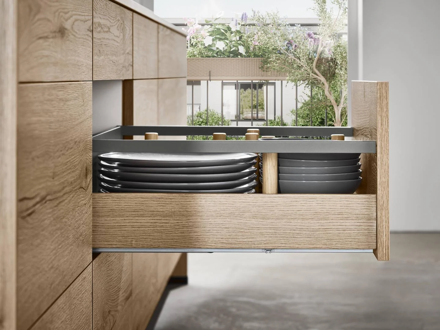 Open drawer containing stacked black plates in a kitchen with wooden cabinetry and a window view of plants outside.