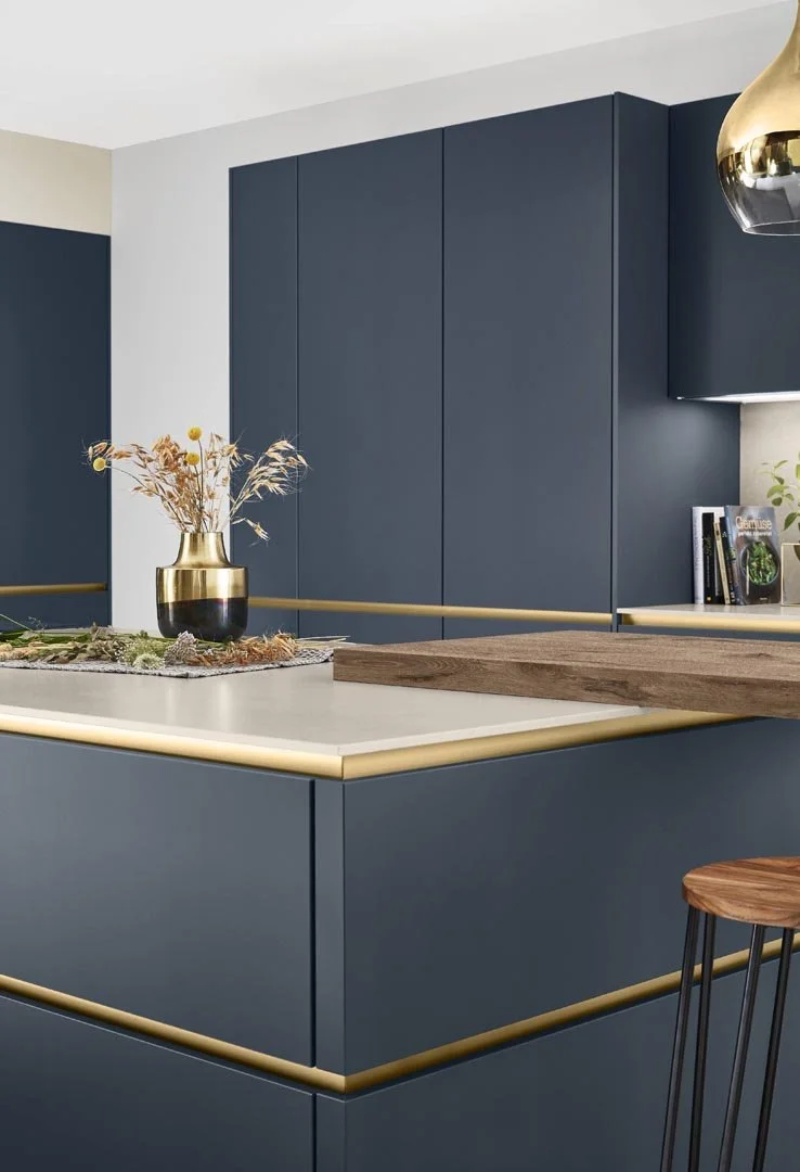 Modern kitchen featuring blue cabinets with gold accents, a white countertop with built-in lighting, a wooden barstool, and decorative items including a gold vase with dried flowers and books.