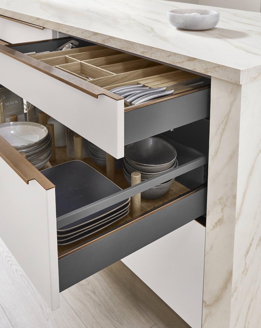 Open kitchen drawer containing wooden utensil dividers and gray and black dishware, with a marble countertop and a small bowl on top.