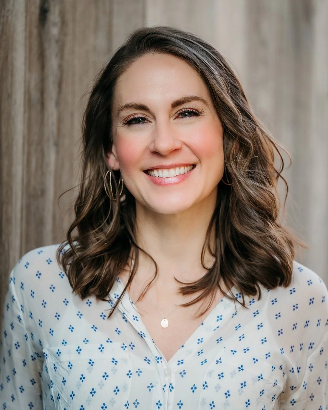 A smiling woman with shoulder-length wavy brown hair, wearing earrings, a necklace, and a white blouse with blue patterns, standing outdoors with a wooden fence in the background.