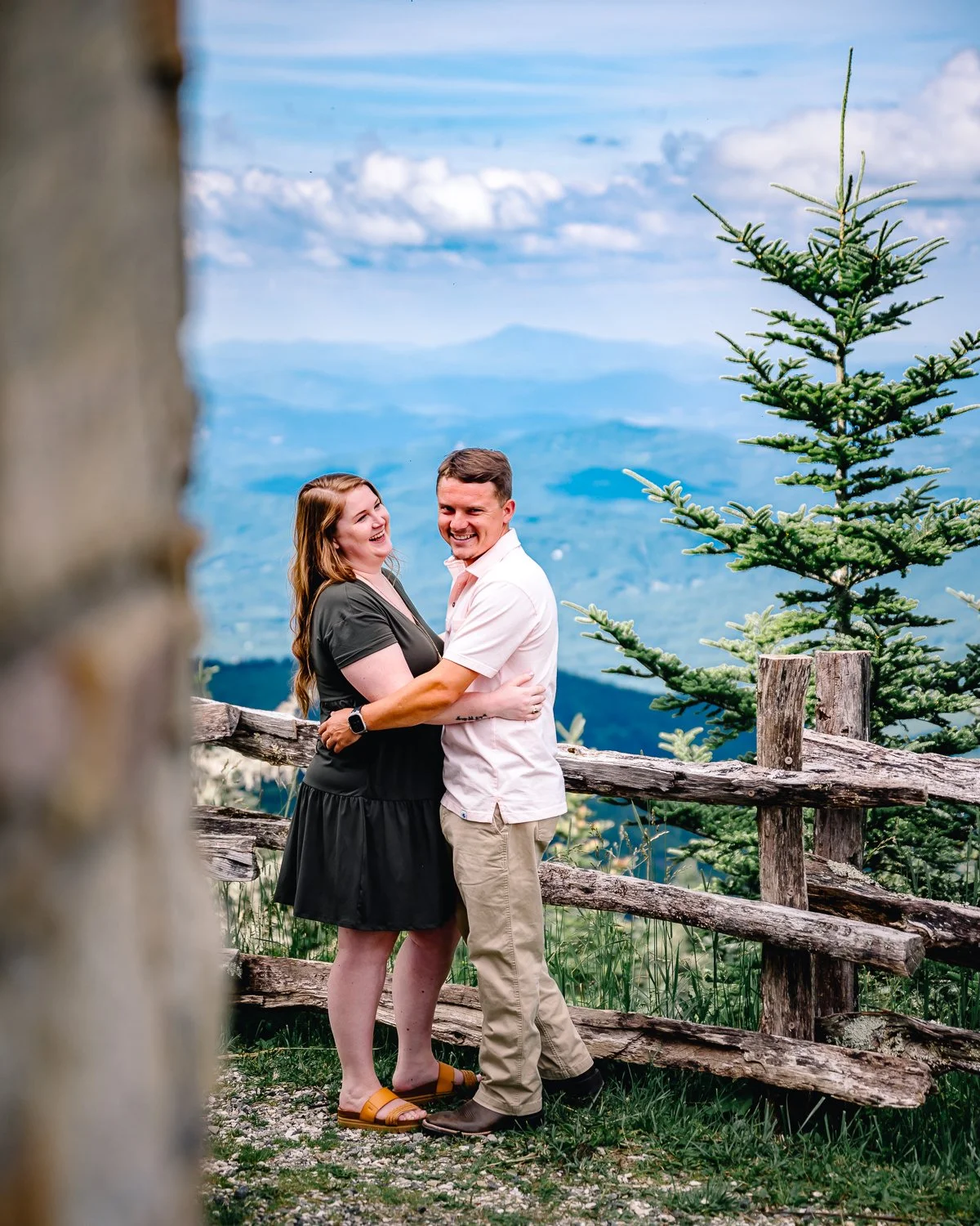 A couple stands outdoors on a grassy area with a rustic wooden fence, embracing and smiling at each other, with a scenic mountain landscape and blue sky with clouds in the background.