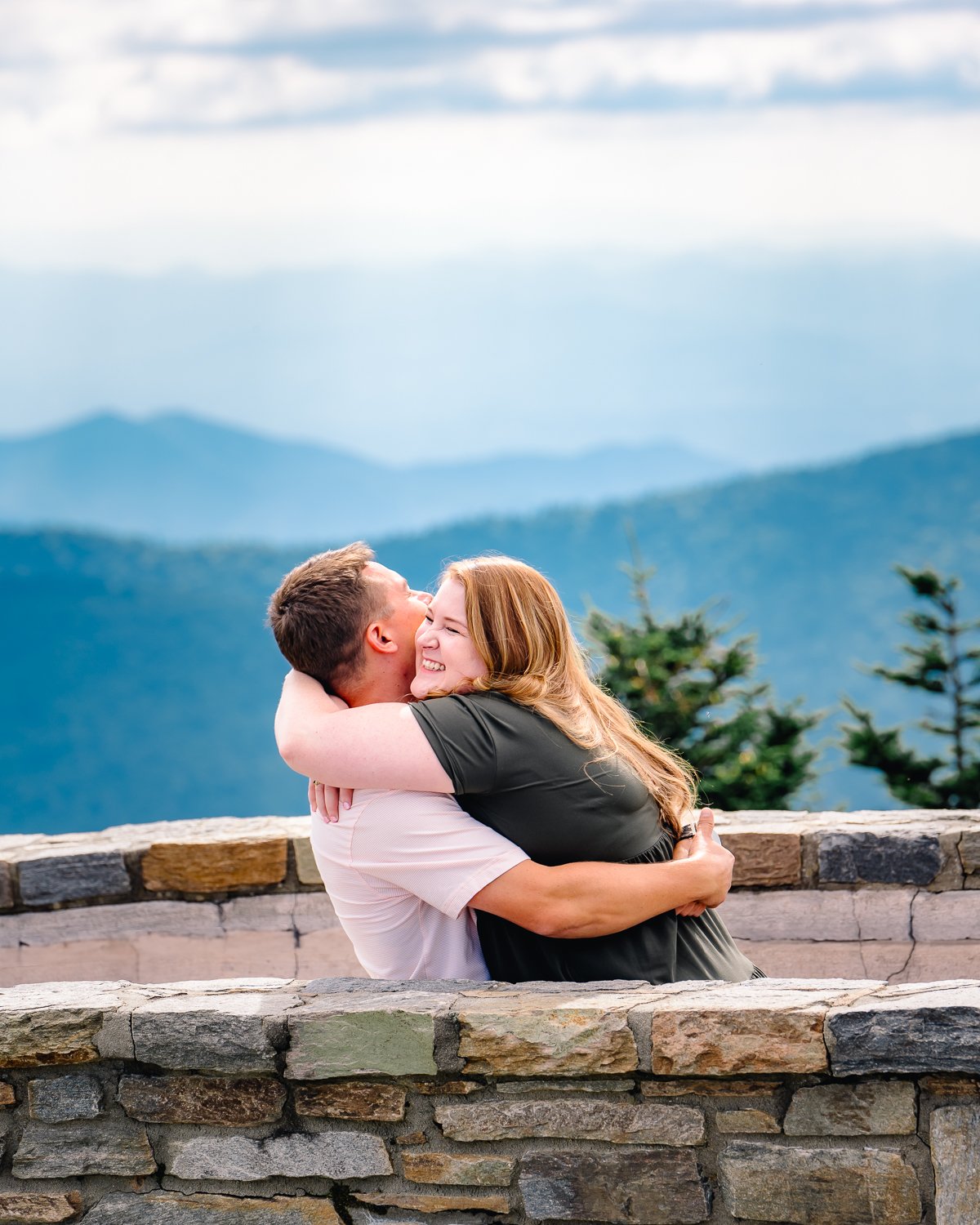 A couple sharing a joyful embrace on a stone wall with mountain views in the background.