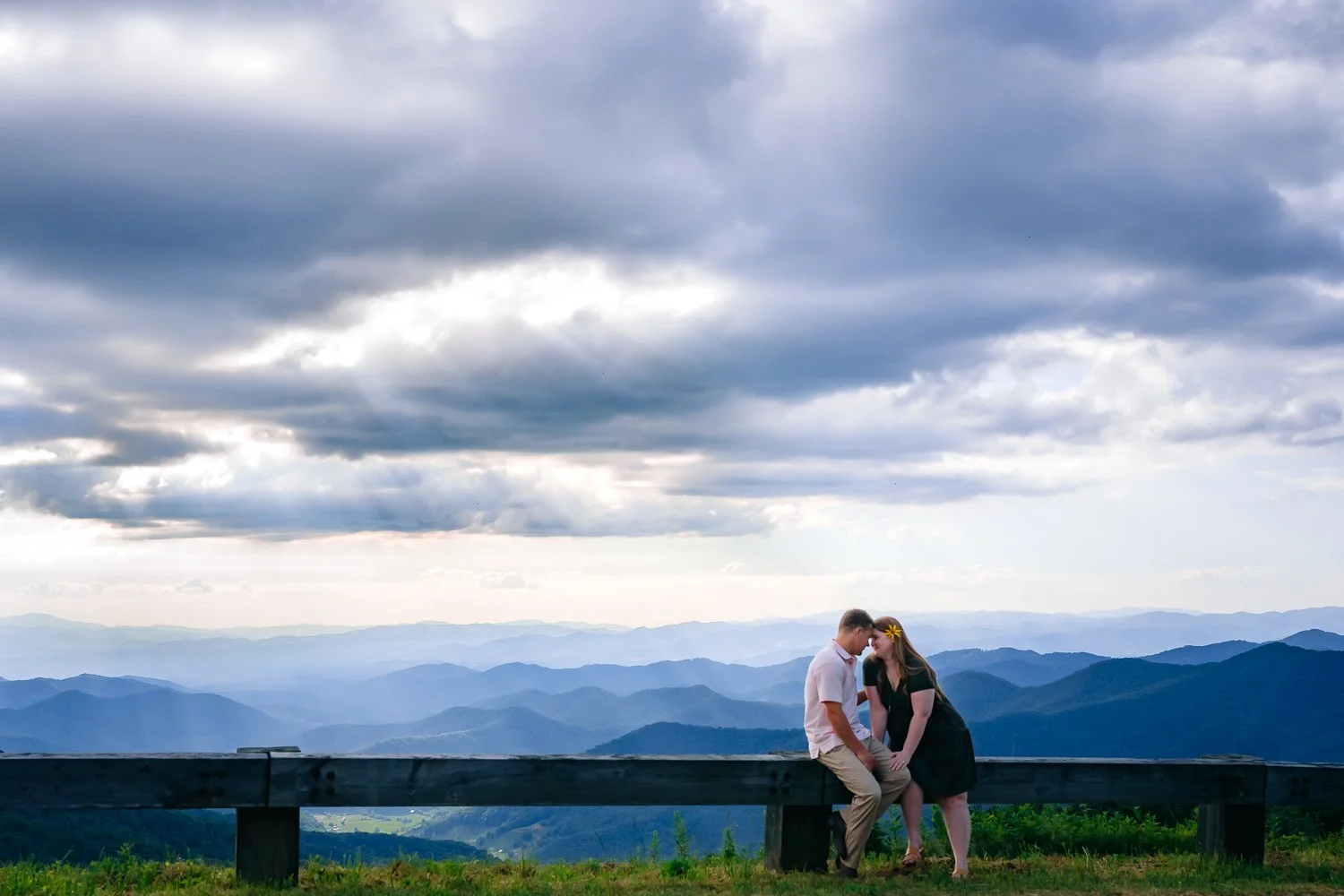 A couple sitting on a wooden railing outdoors with a mountain landscape in the background under a cloudy sky.