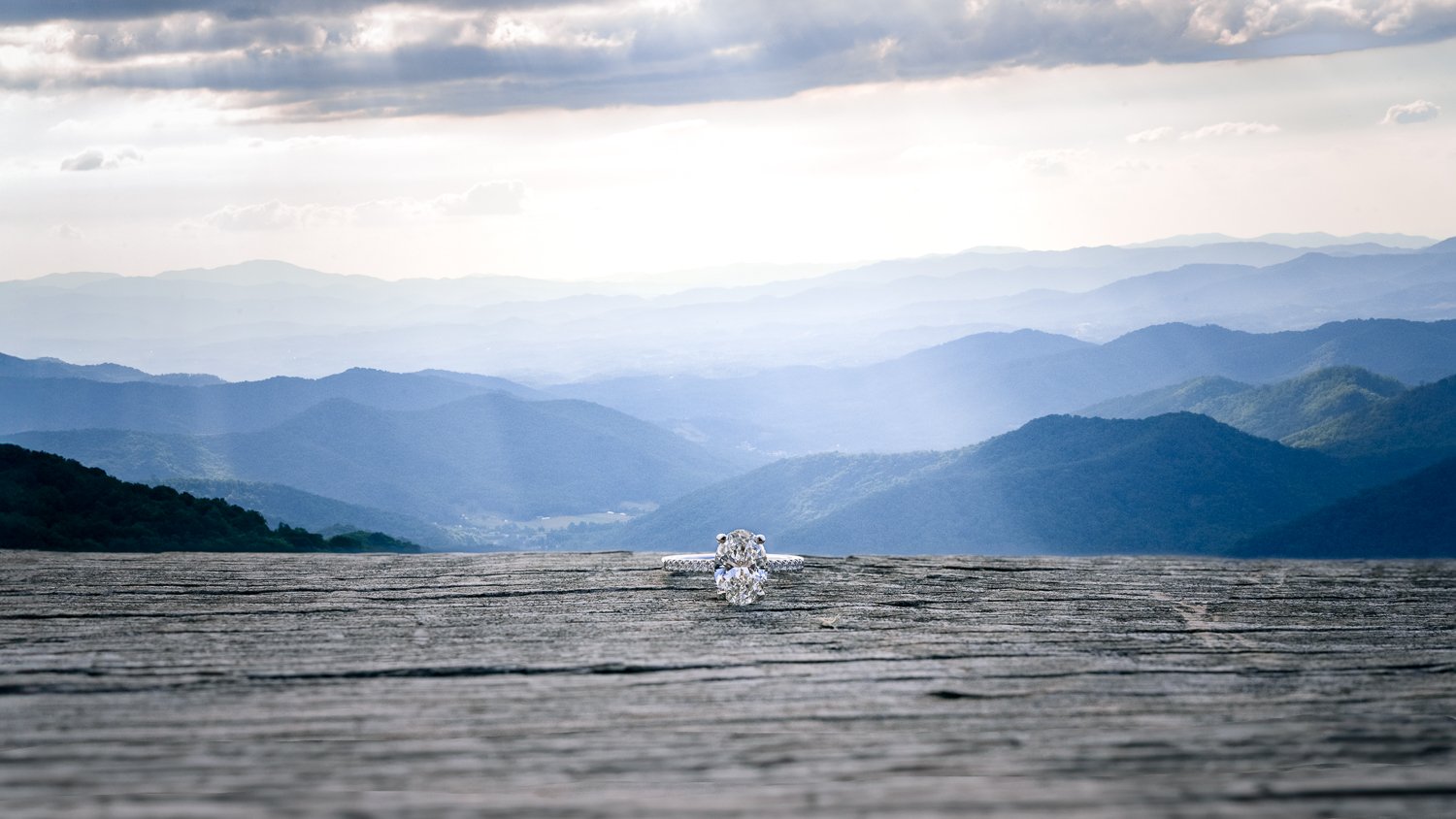 Close-up of a diamond ring on a wooden surface with a scenic mountain landscape in the background, under a cloudy sky.