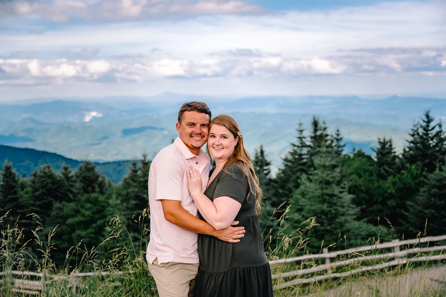 A smiling couple embracing on a hilltop with a backdrop of green pine trees and mountain ranges under a partly cloudy sky.
