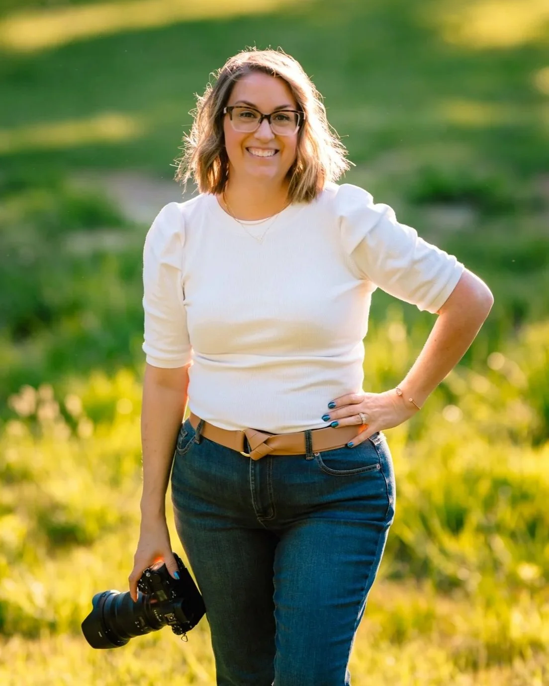 A smiling woman with glasses and shoulder-length hair standing outdoors on a sunny day, holding a camera in her right hand, wearing a white top and blue jeans with a belt, with green grass and trees in the background.