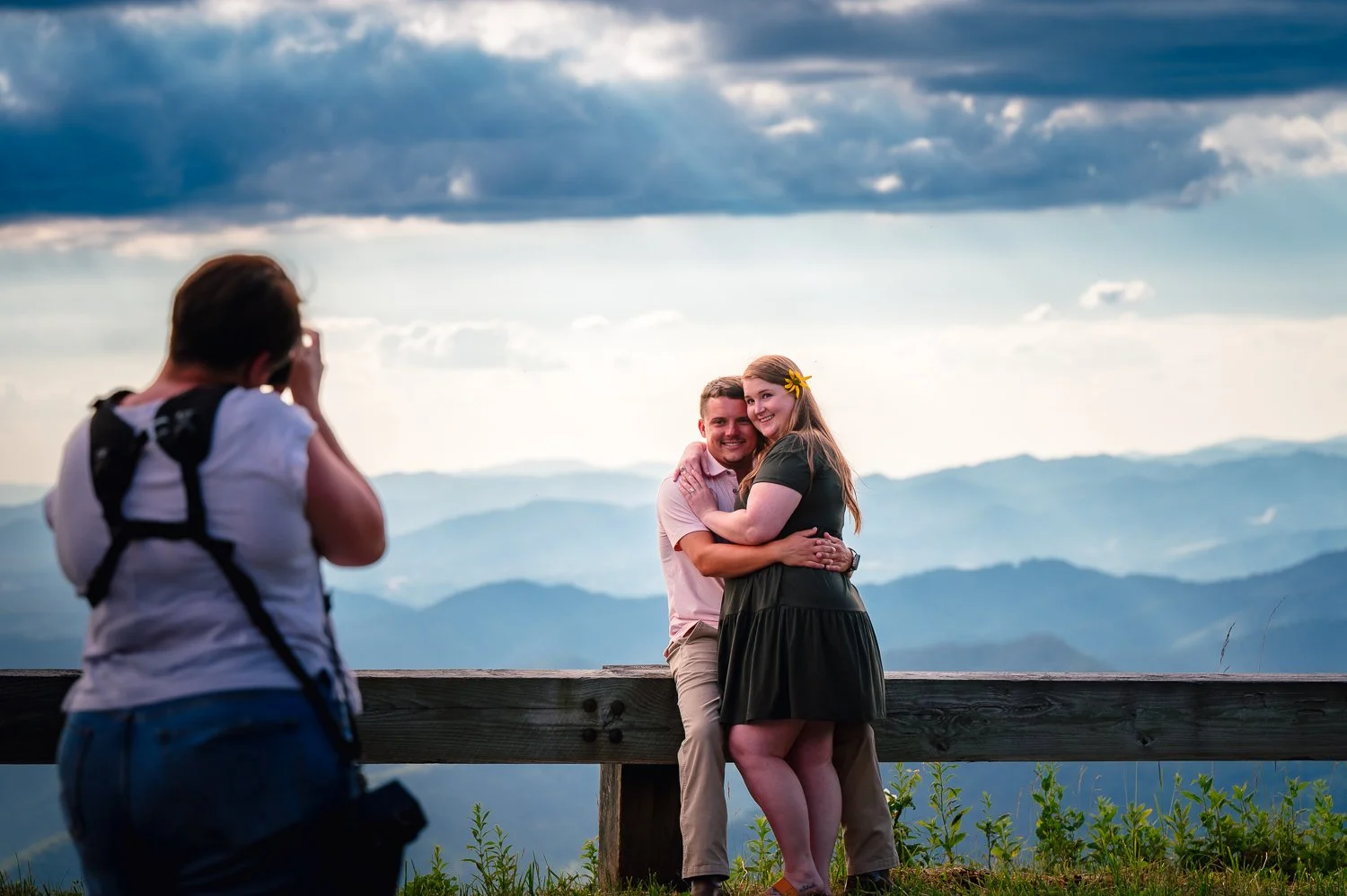A woman and a man sit on a wooden barrier, embracing and smiling, while a photographer takes their picture against a backdrop of mountains and cloudy sky.