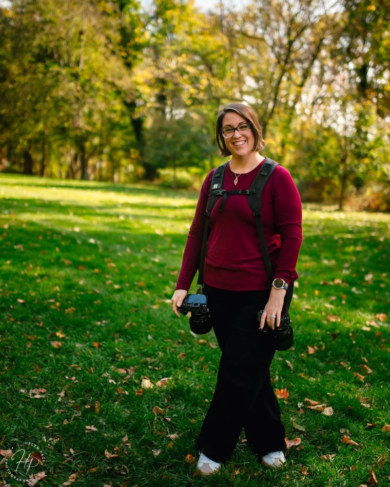 Woman with short brown hair and glasses standing on grass in a park during autumn, holding camera equipment, dressed in a maroon sweater and black pants.