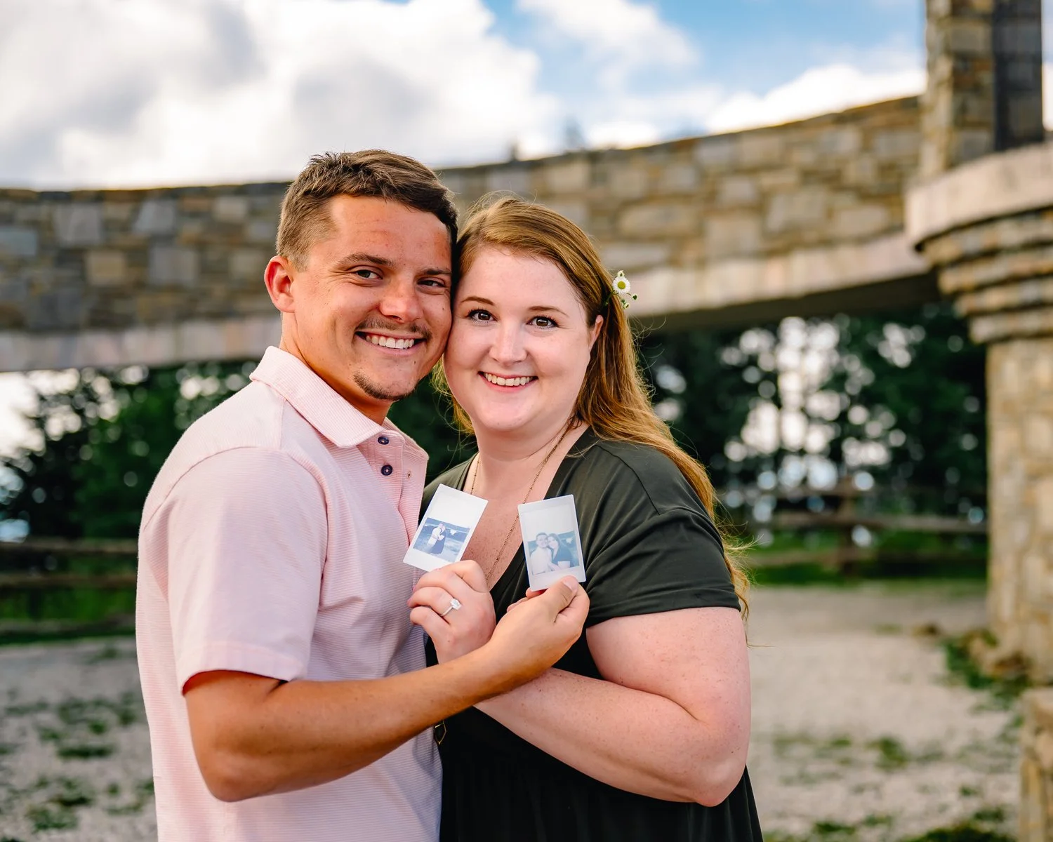 A smiling couple standing outdoors, holding two Polaroid photos, with a stone bridge and trees in the background.
