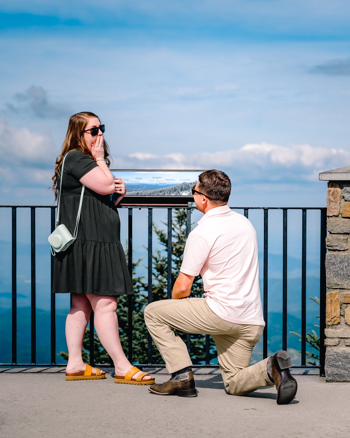 A man kneeling on one knee proposing to a woman on a scenic outdoor observation deck, with a mountain landscape and cloudy sky in the background.