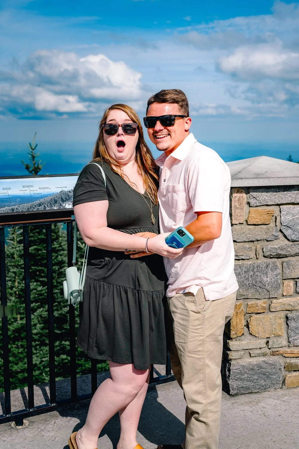 A surprised woman and a smiling man wearing sunglasses at a scenic outdoor viewpoint with a cloudy sky and forested landscape visible in the background.