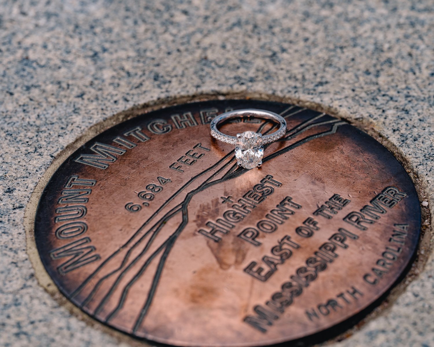 A diamond engagement ring resting on an engraved bronze marker at the highest point east of the Mississippi River atop Mount Mitchell in North Carolina.