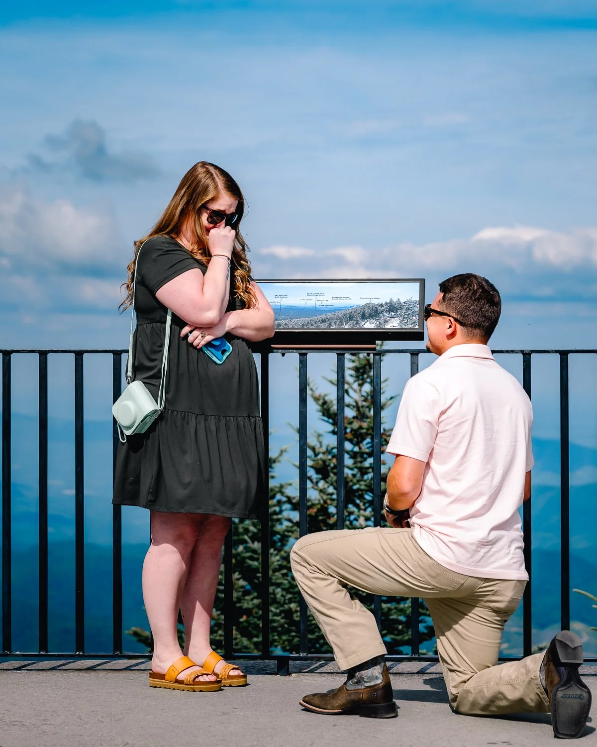 A man is kneeling on one knee proposing to a woman on a scenic overlook with a mountain view and railing. The woman appears surprised, covering her mouth with her hand. The man is looking at her affectionately. There is a sign on the railing with mou