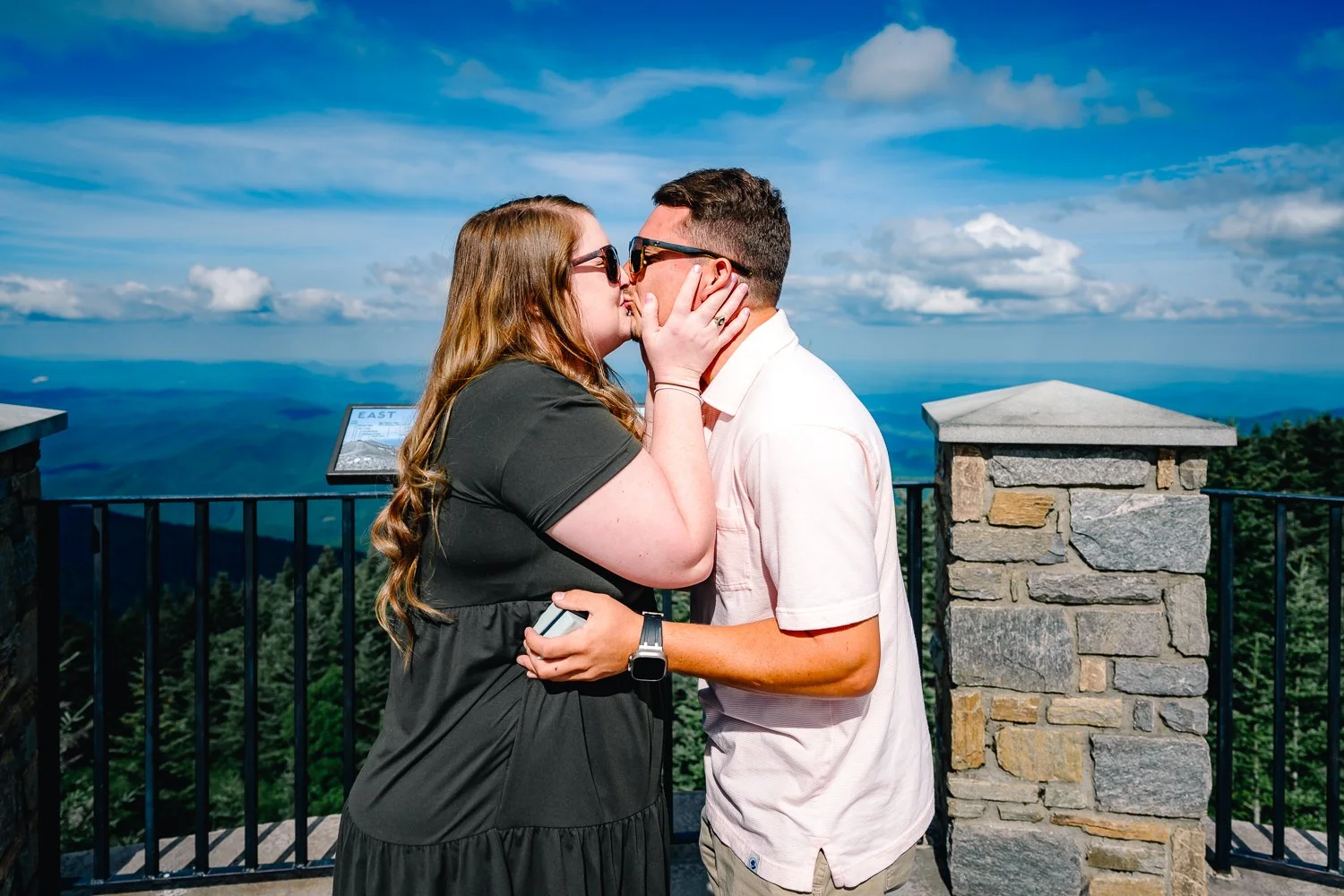 A couple kissing on a scenic overlook with mountains and a cloudy sky in the background.