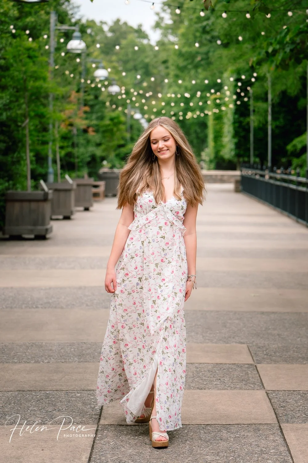 A young woman in a white floral maxi dress walking on a paved path in a park with lush green trees and string lights overhead, smiling and looking downward.