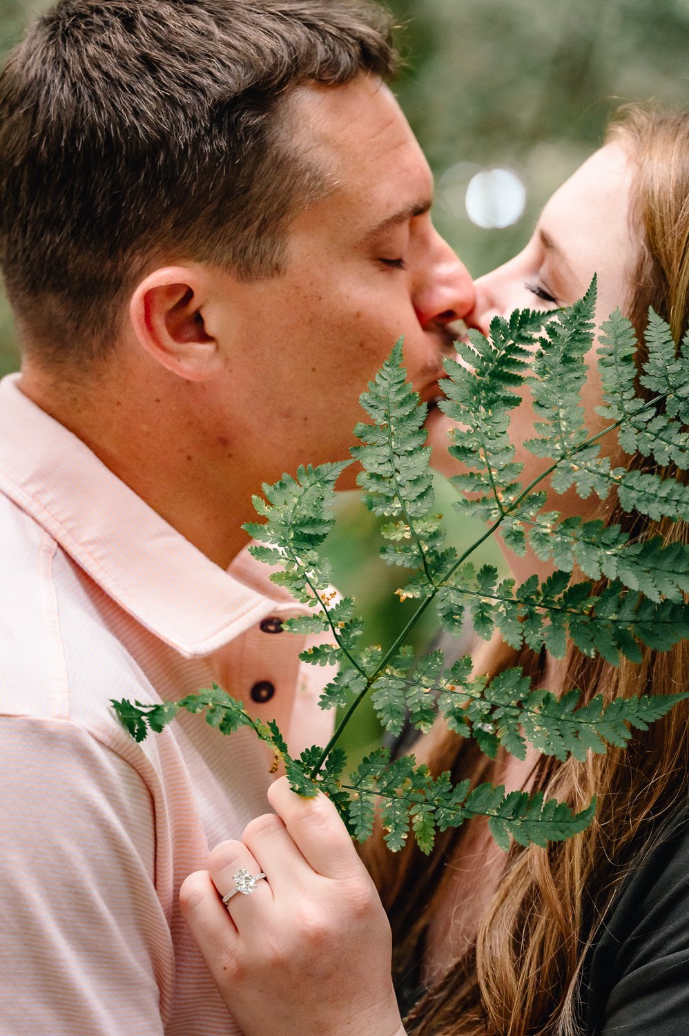 A man and woman sharing a kiss outdoors, with the woman's hand showing an engagement ring and a green fern leaf partially covering their faces.