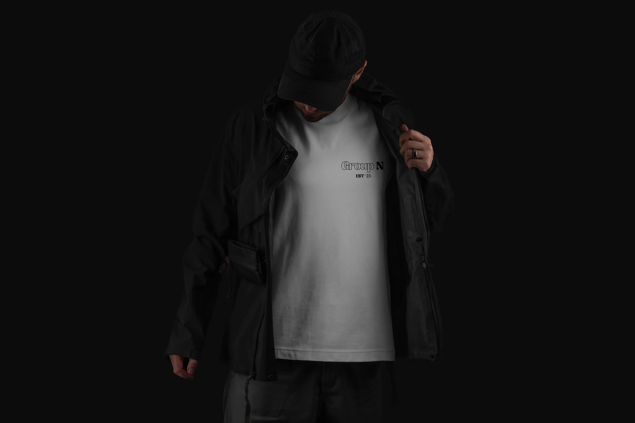 A man wearing a black cap and black jacket, opening his white T-shirt to reveal a design that says 'Group N EST 23'.
