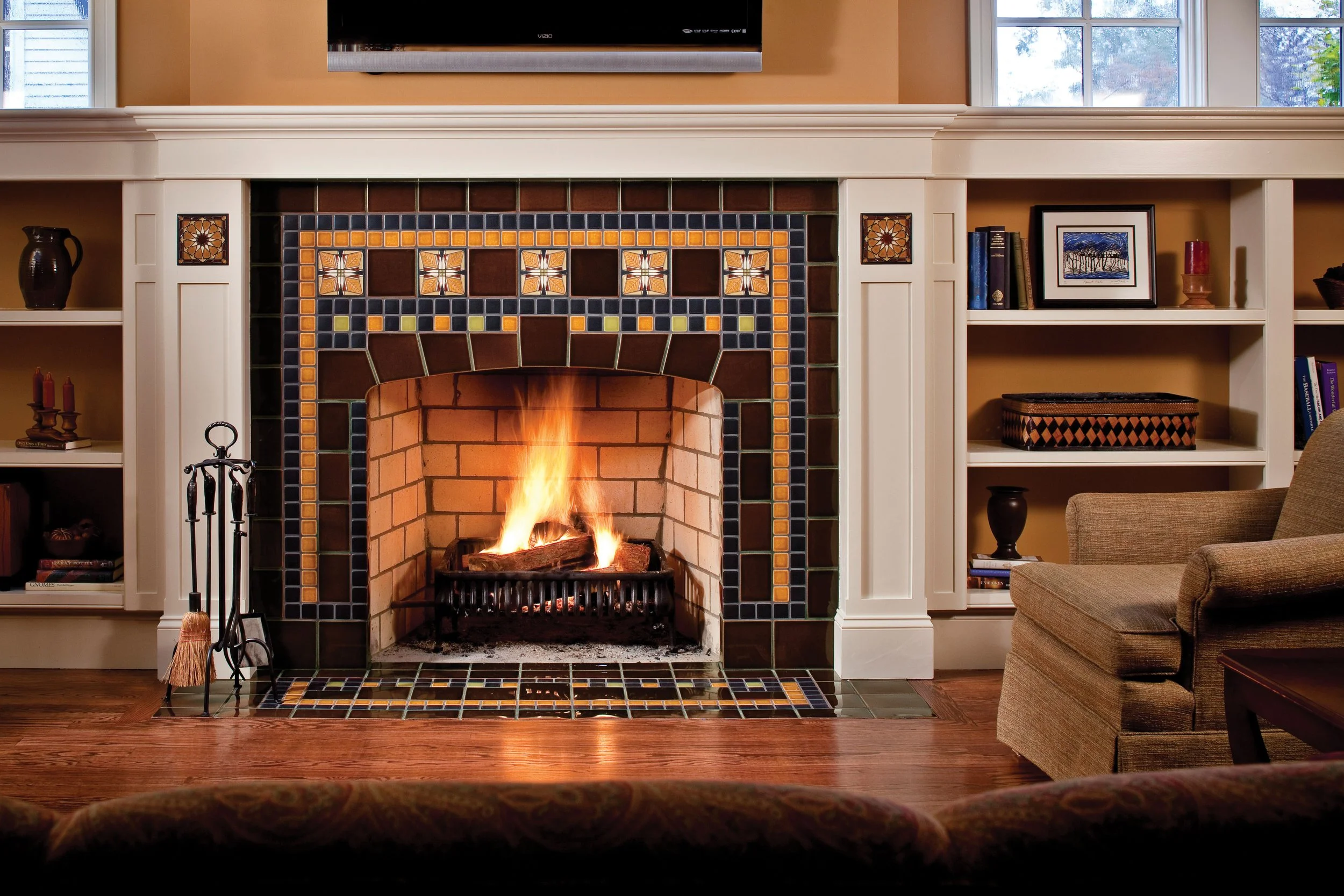 Living room with a lit fireplace surrounded by white built-in shelves, tan walls, hardwood floors, and a beige armchair.