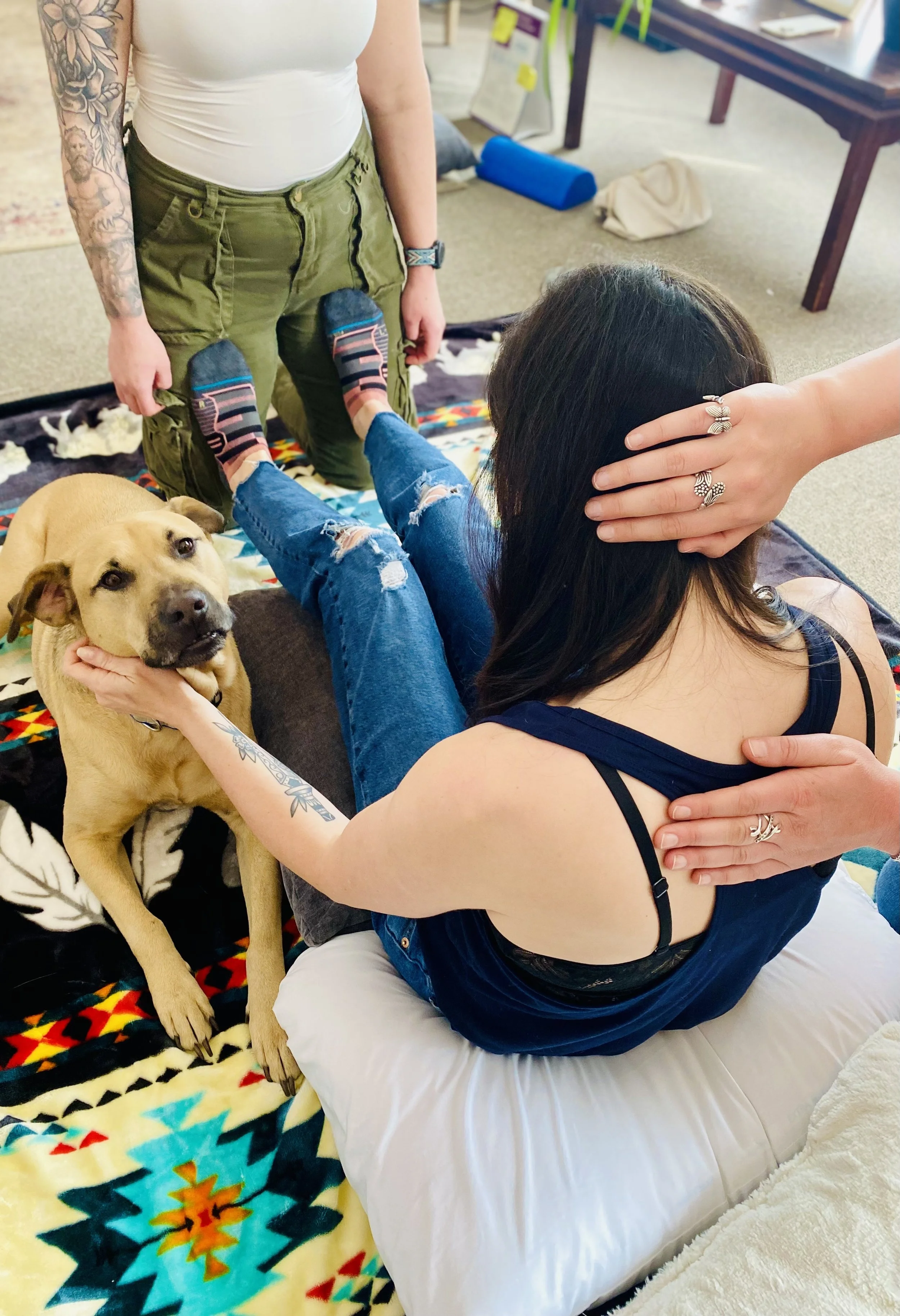 Two women and a dog in a cozy indoor setting. One woman is sitting with her lap supported by another person kneeling, while the dog rests its head on her shoulder and looks at the camera. The women are touching each other's heads and arms, and the scene is relaxed with colorful blankets and a casual office or living room background.