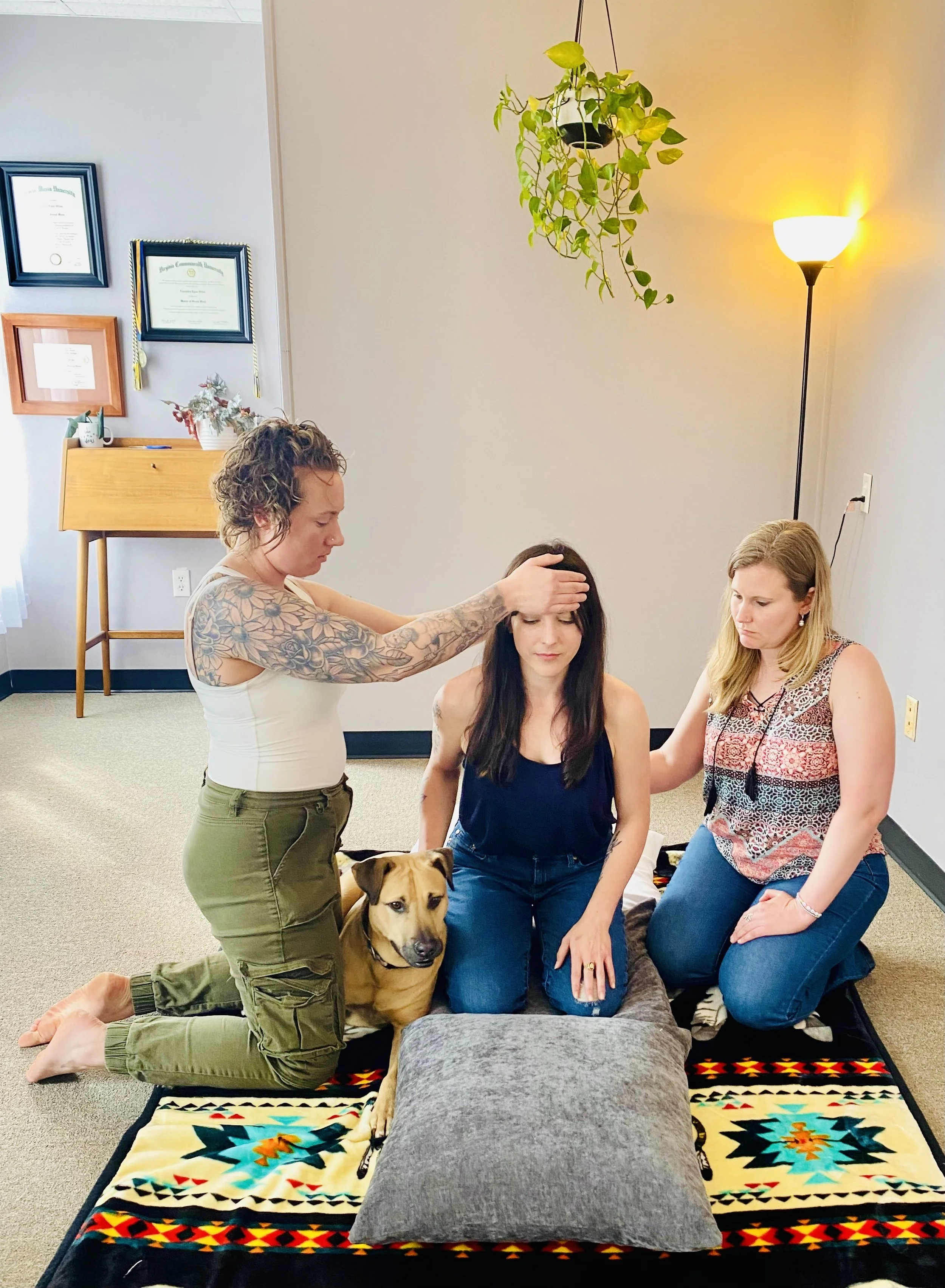 A woman receives a healing energy session with two women and a dog in a sitting meditation posture in a room with certificates on the wall, a hanging plant, and a standing lamp.