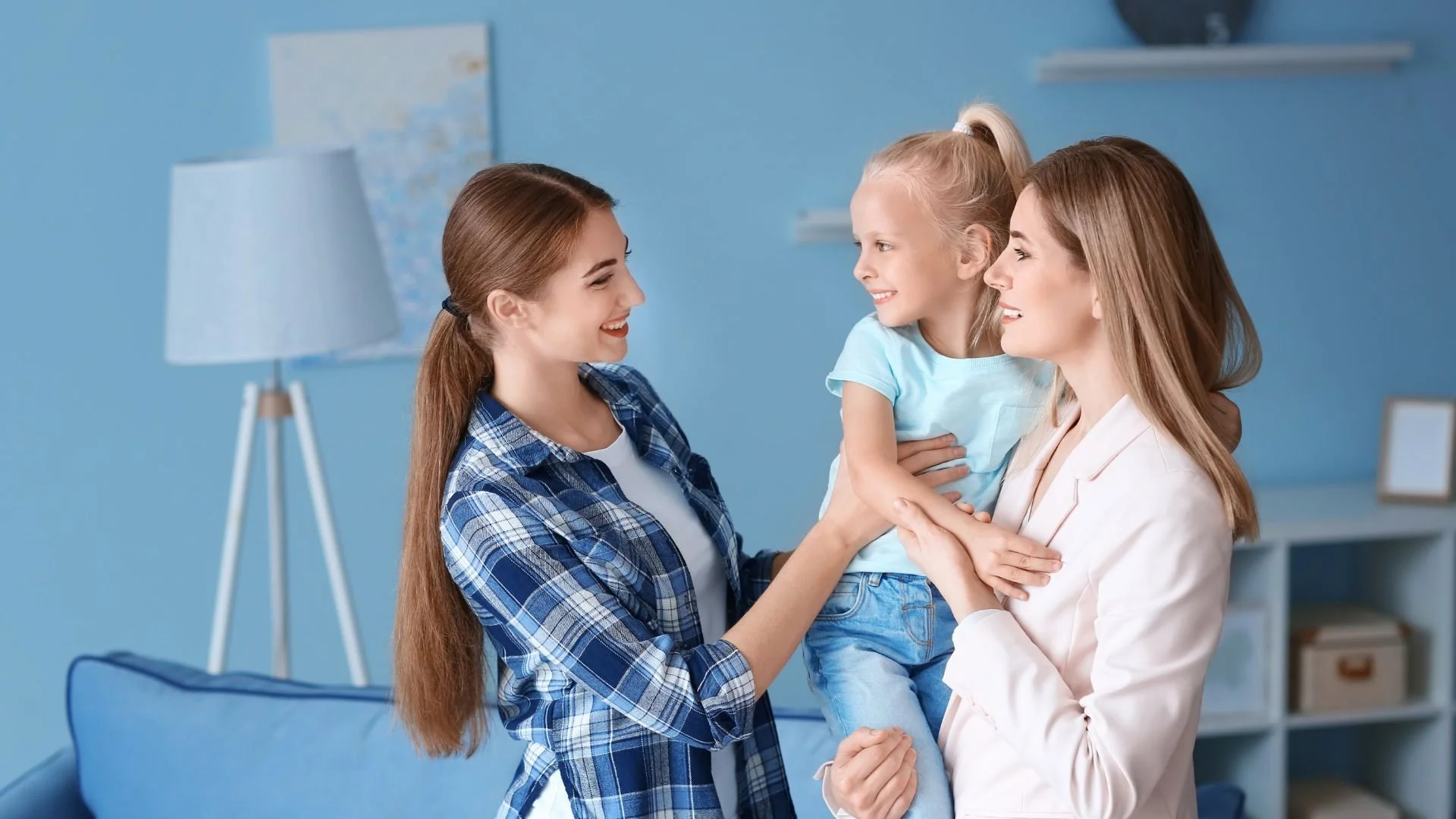 A smiling mother comfortably handing over her toddler daughter to a professional AMP Placement nanny at the front door of a modern home.