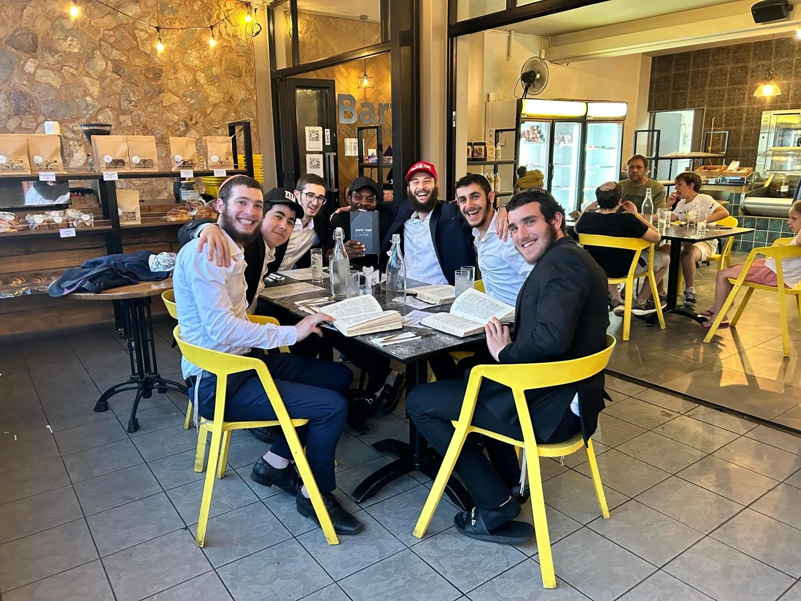 Group of men sitting around a table reading books in a café with a rustic interior design, featuring yellow chairs and shelves of baked goods.