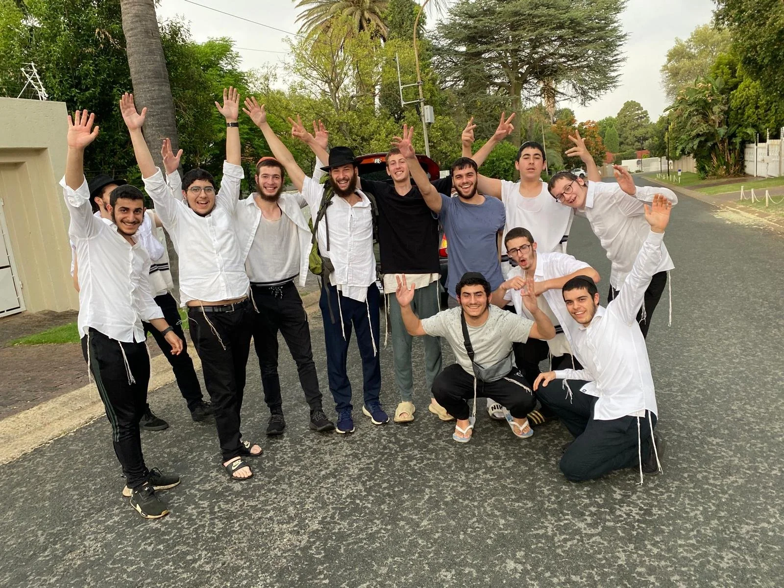 Group of young men standing on a street, smiling and raising their hands cheerfully, with greenery and a palm tree in the background.
