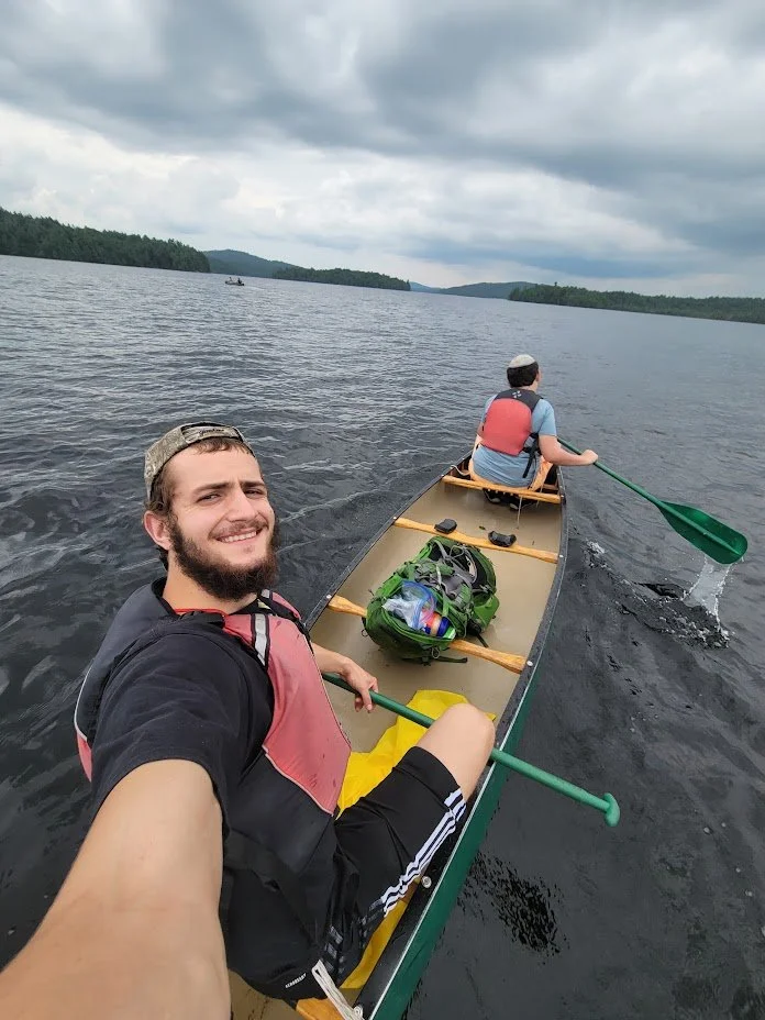 Two men canoeing on a lake under cloudy skies, with one taking a selfie. They are wearing life vests, with a backpack visible in the canoe.
