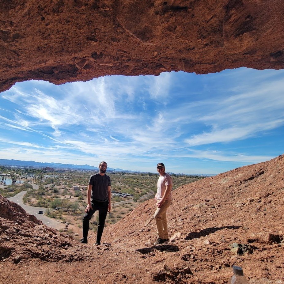 Two people standing in a rocky landscape under a natural rock arch with a clear blue sky and distant mountains in the background.