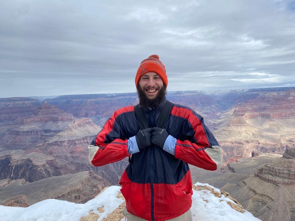 Person in a red and blue jacket standing at the edge of the Grand Canyon, smiling with snow on the ground and a cloudy sky.
