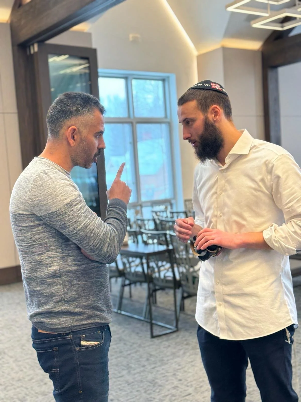 Two men standing indoors having a conversation near a window; one man with a beard wearing a kippah and holding headphones.