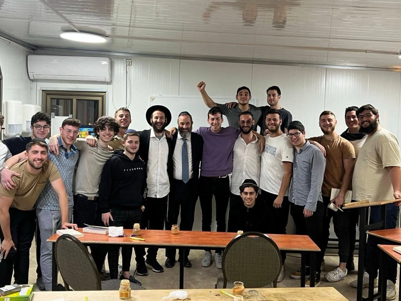 Group of men posing indoors, some wearing hats, in a room with tables and chairs.