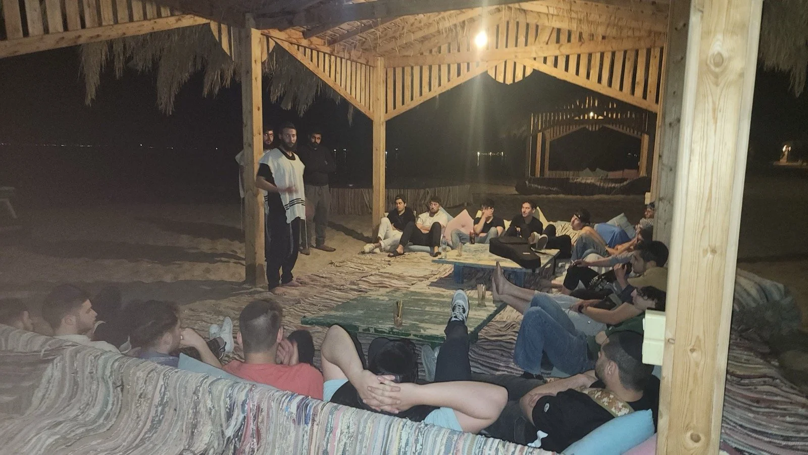 Group of people sitting under a wooden canopy at night, some on couches and some standing, engaged in conversation or listening.