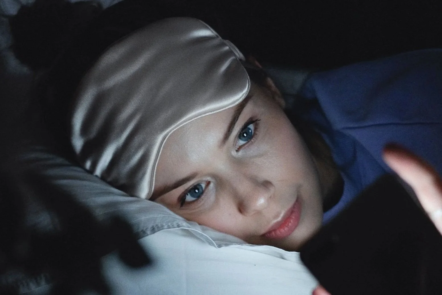 Women laying in bed in the dark with a sleep mask on her forehead, looking at her phone. The phone screen is illuminating her face.