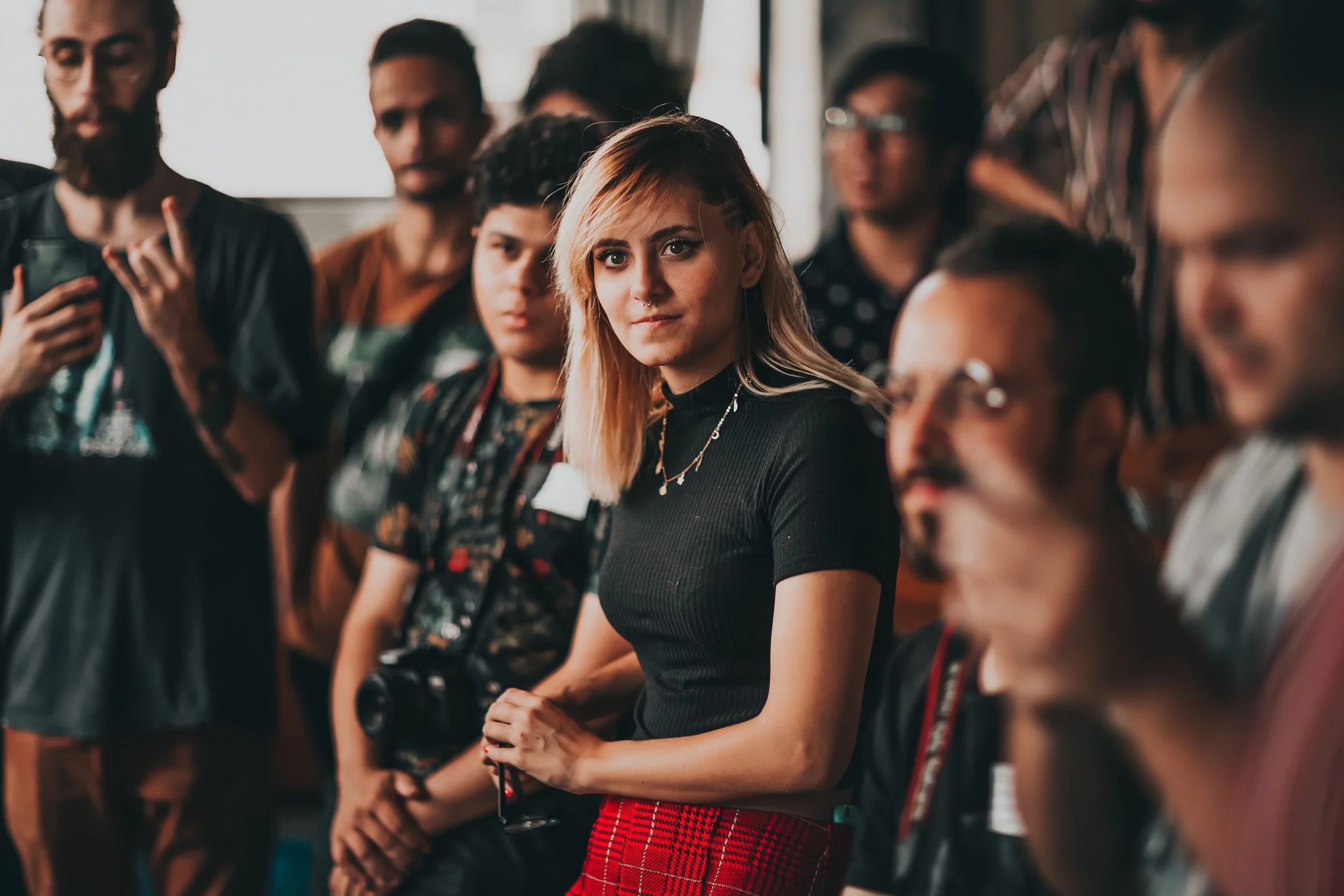 Picture of a woman standing in a crowd of other people that are blurry. She has a camera in her hands and is intently looking at something. She has long, blonde hair with a shaved section above her ear. She has a nose ring, wearing a black shirt.