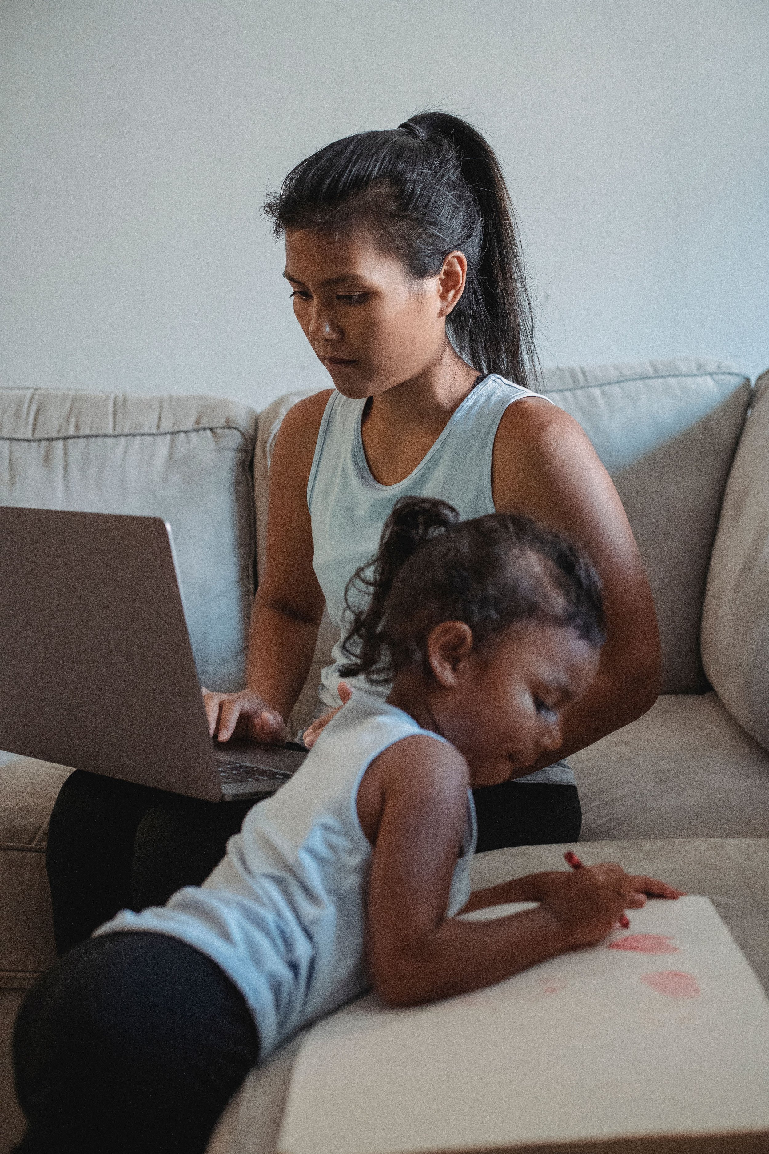 Woman and her daughter at home together. Woman is wearing a light blue sleeveless shirt with dark leggings, sitting on a beige couch with her laptop on her legs. She has dark hair in a ponytail. Her daughter is dressed similarly, standing , drawing.