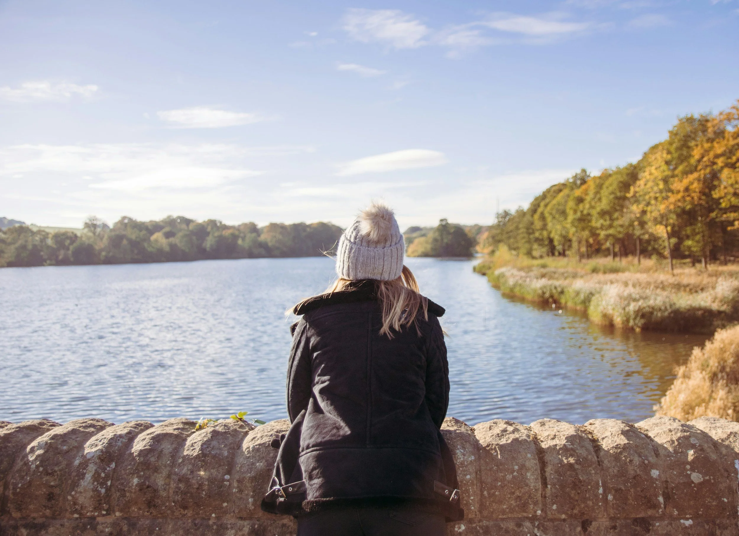 A woman wearing a gray beanie and black jacket looking at a lake with trees showing autumn colors.
