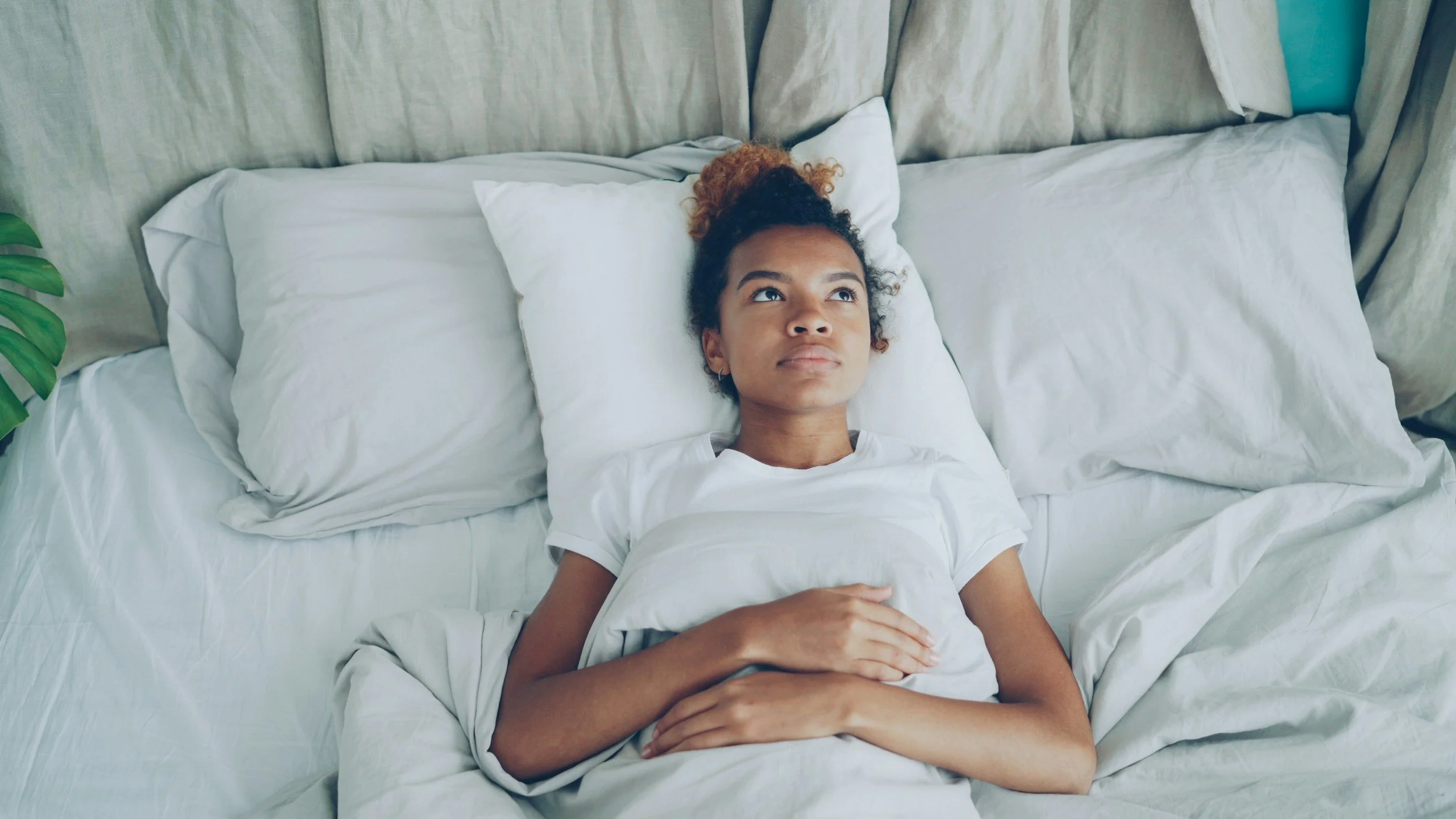 A young woman lying in bed looking up at the ceiling with a neutral expression. She has curly hair tied up and is covered with white sheets. The bed has white pillows, and a green plant is partially visible on the left side of the image.